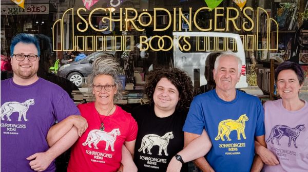 Row of smiling people who have lined arms wearing purple, red, black, blue and yellow t-shirts.
