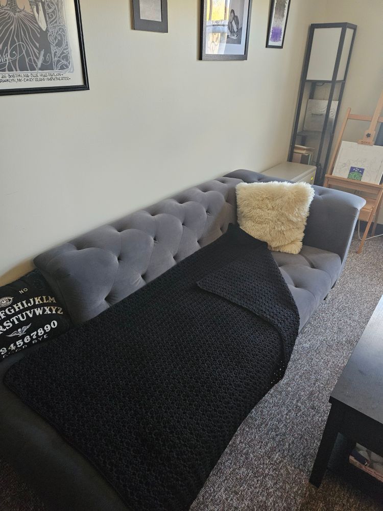 A black wool couch-length blanket crocheted in a modified shell stitch on a grey chaise lounge. There is also a sheepskin pillow and a ouiji board neck pillow.