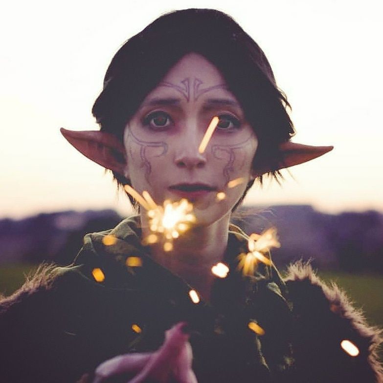 A cosplayer dressed as Merrill from Dragon Age 2 extends her hand to the camera at twilight, sparks flying from her fingers. Red light reflects in her eyes, she has a neutral expression 