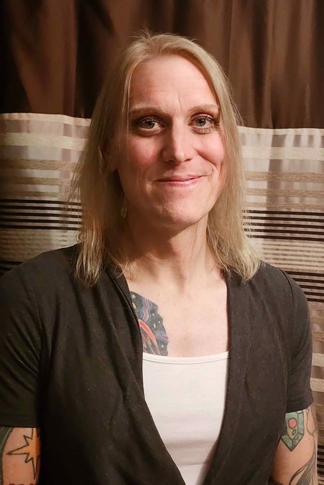 A photo of me (trans woman with blonde hair, tattoos on her arms and chest), wearing makeup for the first time, in front of a shower curtain