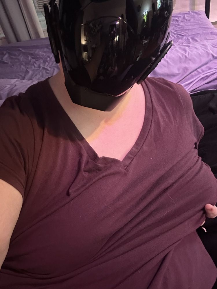 A drone girl in a red shirt wearing a featureless black visor stares into the camera. She's laying against a soft mattress with lavender sheets.