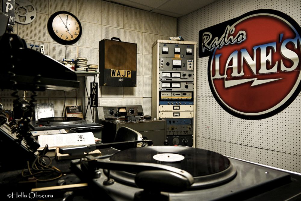 a photo of a replica of the old radio station WAJP in Joliet, IL. Sound dampening panels, dingy with age, line the walls. "Radio Lane's" sign adorns the back wall. A large turntable is directly in front of the camera, a telephone on a shelf to the left. A portable speaker hangs on the far wall, above a reel-to-reel. Tapes sit on a shelf above another turntable, and a tall cabinet stands in the far corner housing all sorts of the electrical and musical devices needed for broadcasting. 
