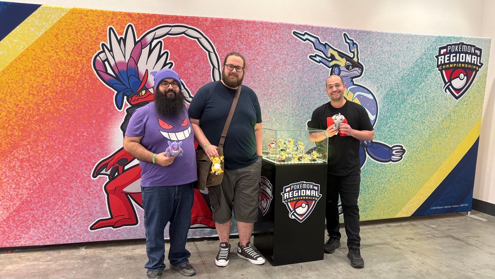 A picture of @chrispynintendo, @galutrad, and Aaron standing in front of a banner featuring Koraidon and Miraidon and the “Pokémon Regional Championships” logo. Between Aaron & @galutrad is a glass case featuring the medals that the top players received. 

@chrispynintendo is holding a Gengar plush, @galutrad has Raichu, and Aaron has Espurr