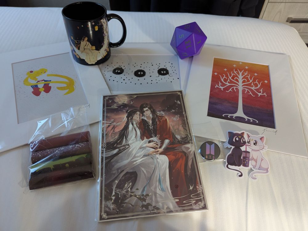 Photo showing a TGCF mug and print, a Sailor Moon art, a Gondor tree art, a soot sprites art, a purple 20 sided die stress ball, a sticker with Luna and Artemis, a BTS logo pin, and 4 pieces of chocolate 