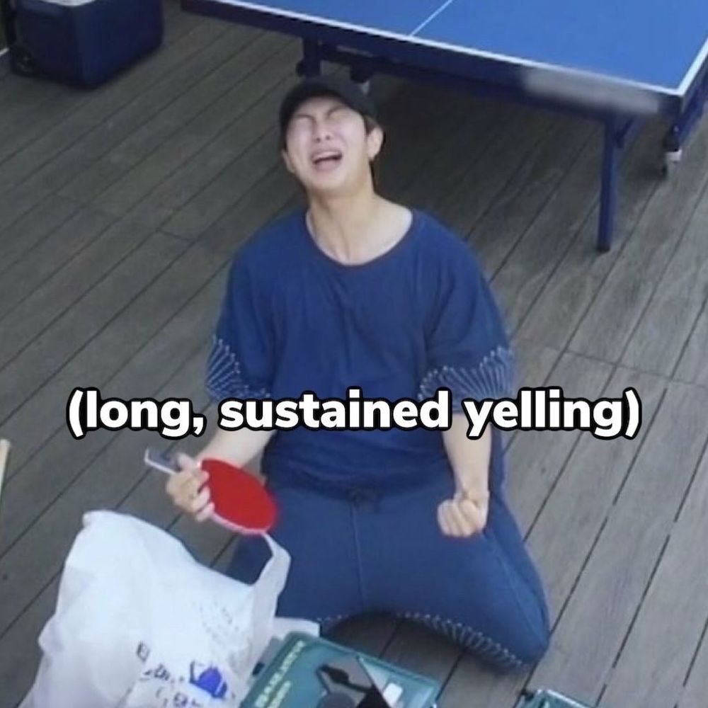 Kim Namjoon wearing blue and on his knees with a red ping pong paddle in his hands. His head is thrown back and he's screaming. It's captioned (long, sustained yelling)