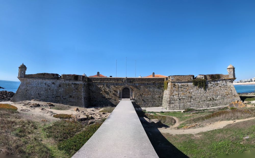 Panoramic view of Forte de São Francisco Xavier, also known as Castelo do Queijo, a historic stone fort on the coast of Porto, Portugal. Th photo shows the fortress’s solid granite walls, corner watchtowers, and arched main gate under a clear blue sky, with the Atlantic Ocean visible to the left near the surf school on Porto beach.