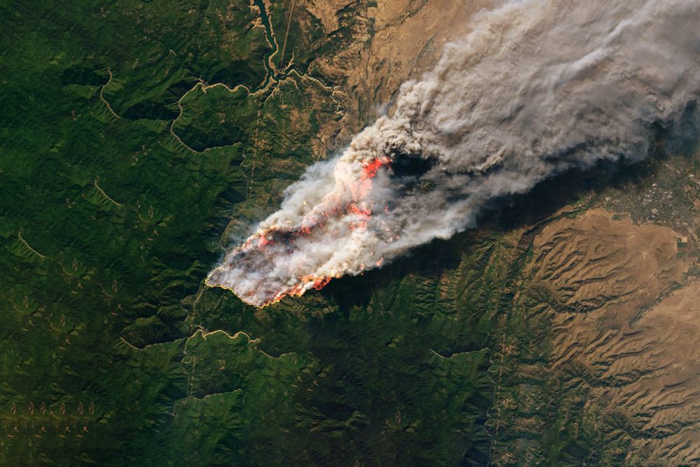 Satellite view of an intense wildfire burning through forested land, with thick smoke spreading toward a populated area.