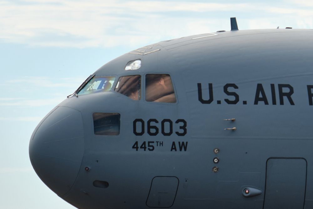 Close-up of the cockpit and front fuselage of a grey U.S. Air Force C-17 Globemaster III military transport aircraft. The numbers '0603' and '445TH AW' are visible below the cockpit windows. Someone in the cockpit waving is visible.