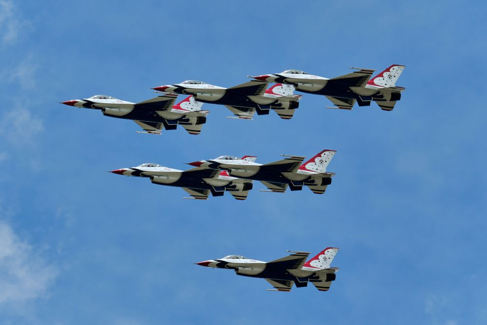 A group of F-16 Fighting Falcon jets, painted in the distinctive red, white, and blue colors of the United States Air Force Thunderbirds, fly in a tight formation against a blue sky with some wispy white clouds.