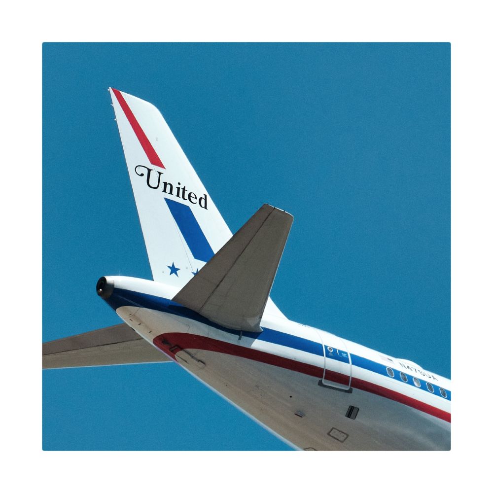 A low-angle shot captures the tail section of a United Airlines A320 airplane against a clear, vibrant blue sky. The plane's white fuselage is visible, adorned with a red,white, and blue cheat line that runs along its side. The prominent tail fin is also white, featuring the "United" logo in black script near the top within a stylized red and blue stripe. A white border frames the shot.