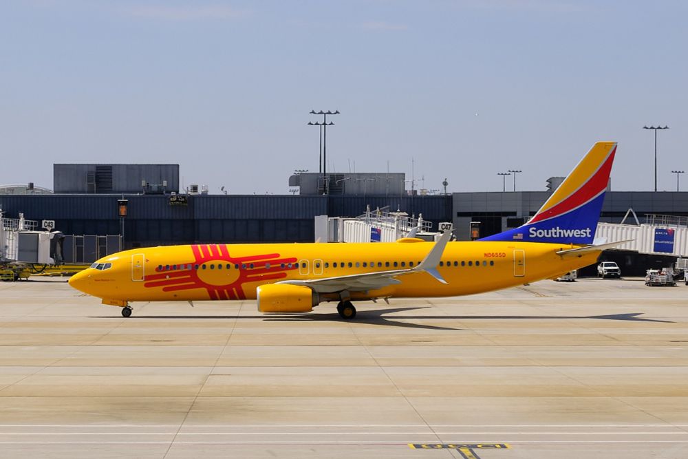 A vibrant yellow Southwest Airlines Boeing 737, tail number N8655D, is parked on an airport tarmac under a clear blue sky. The aircraft features a large red and orange Zia sun symbol on its fuselage, part of a special livery. The tail of the plane is blue with red and yellow stripes, displaying the Southwest Airlines logo. In the background, airport buildings and jet bridges are visible, along with some ground support equipment. The overall impression is a bright and sunny day at the airport.