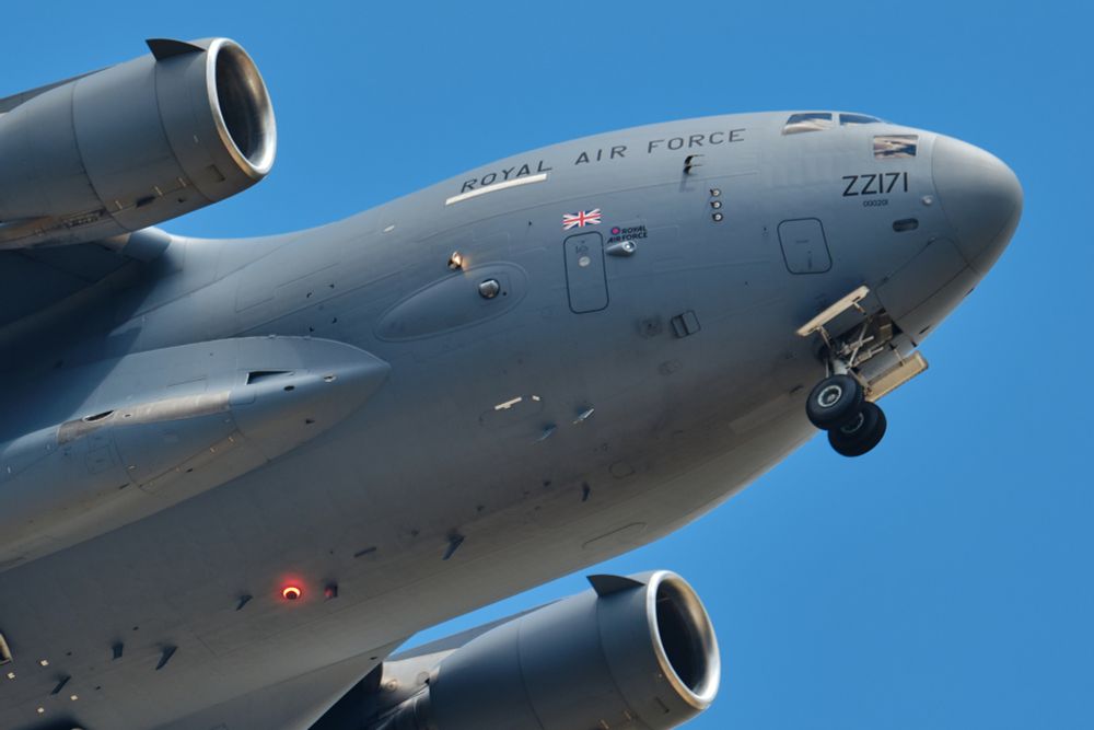 A close-up, low-angle shot of the front and underside of a Royal Air Force C-17 Globemaster III military transport aircraft, registration ZZ171, in flight against a clear blue sky. Its landing gear is deployed. The aircraft's engines and the "ROYAL AIR FORCE" text are visible.