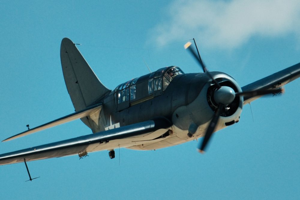 A low-angle, full shot shows aSB2C Helldiver aircraft in flight against a bright blue sky with scattered white clouds. The aircraft is painted in a two-tone blue and white camouflage, with the white on the underside. The US white star insignia is visible on the side of the fuselage and on the top of the right wing. The plane is banking to the right, and the propeller is blurred from motion.