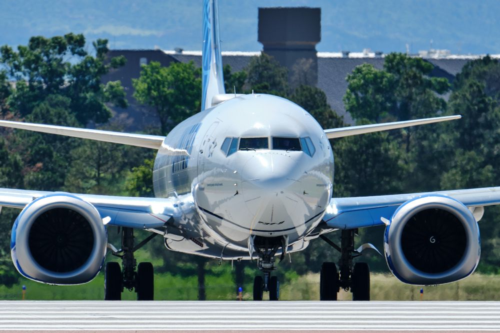  A United Airlines Boeing 737 MAX 9, registration N37536, is shown head-on, with its nose and cockpit prominent. The aircraft's jet engines are visible on either side. In the background, dark green trees and a building are partially visible under a bright sky. The aircraft appears to be on a paved surface.
