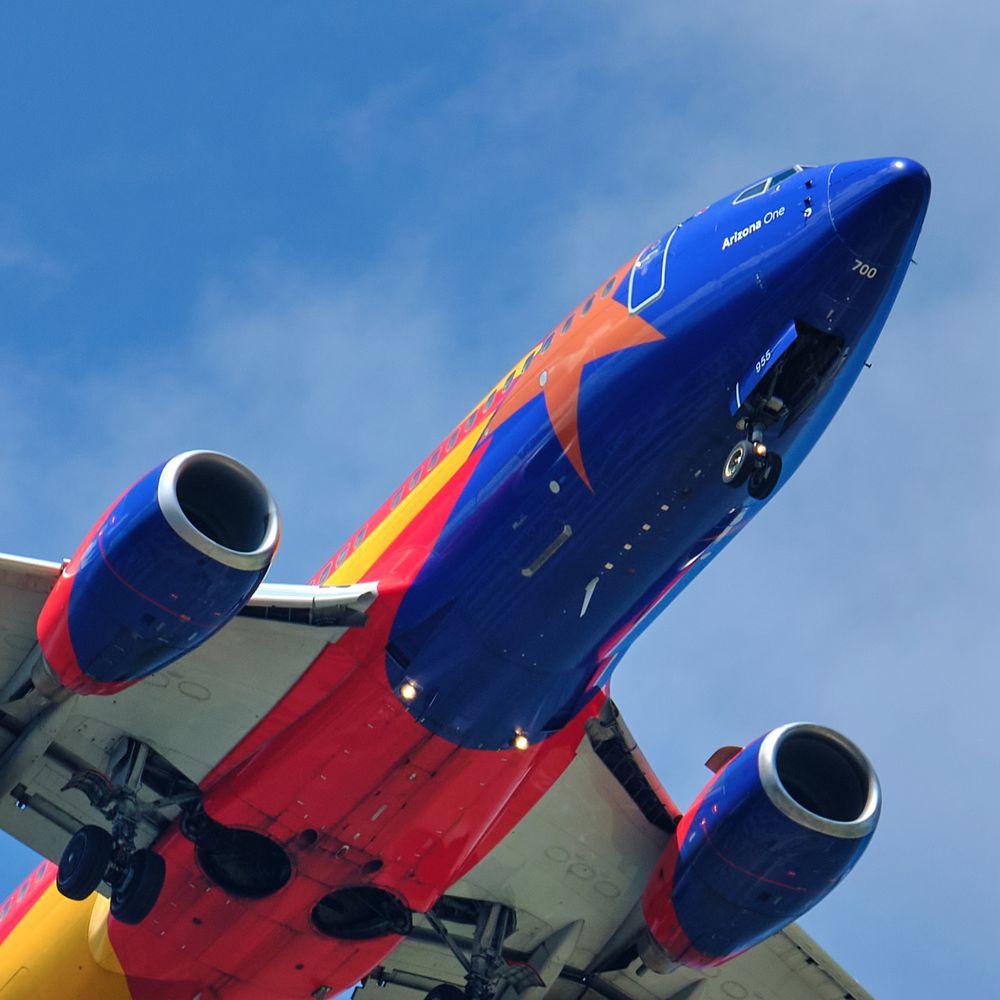 A low-angle shot captures the underside of a Southwest Airlines Boeing 737, specifically the "Arizona One" special livery, against a bright blue sky with scattered clouds. The aircraft's fuselage is primarily dark blue on top, transitioning to red and yellow stripes along its belly, with a large orange star decal visible near the front. The two engine nacelles are also blue and red. The landing gear is extended, showing its wheels and struts.