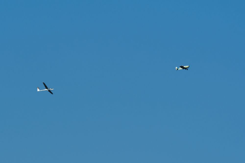 A white DG-1000S glider soars on the left side of a clear blue sky, while a small, light-colored tow plane with green and blue markings is visible further to the right. A barely visible cable connects both planes.