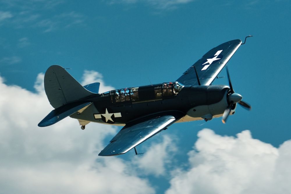 A low-angle, full shot shows a SB2C Helldiver aircraft in flight against a bright blue sky. The aircraft is painted in a two-tone blue and white camouflage, with the white on the underside. The US white star insignia is visible on the side of the fuselage and on the top of the right wing. The plane is angled slightly upwards, and the propeller is blurred from motion.