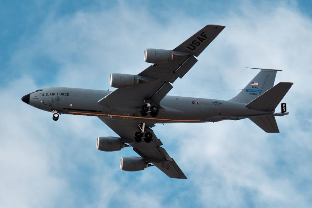 A U.S. Air Force KC-135 Stratotanker is captured from a low, almost directly beneath angle as it flies overhead with its landing gear deployed. The matte gray fuselage, refueling boom housing, and four underwing engines are all sharply defined. “U.S. AIR FORCE” markings, tail number, and the Air Force Reserve Command tail band are visible. The aircraft is framed against a soft blue sky streaked with thin, diffuse clouds, giving the tanker a dramatic, looming presence above.