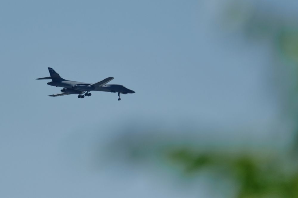 A dark grey B-1B Lancer bomber aircraft with its landing gear down  descends against a slightly smoky blue sky. The aircraft is angled slightly upward from left to right, with faint blurred green foliage in the bottom right foreground.
