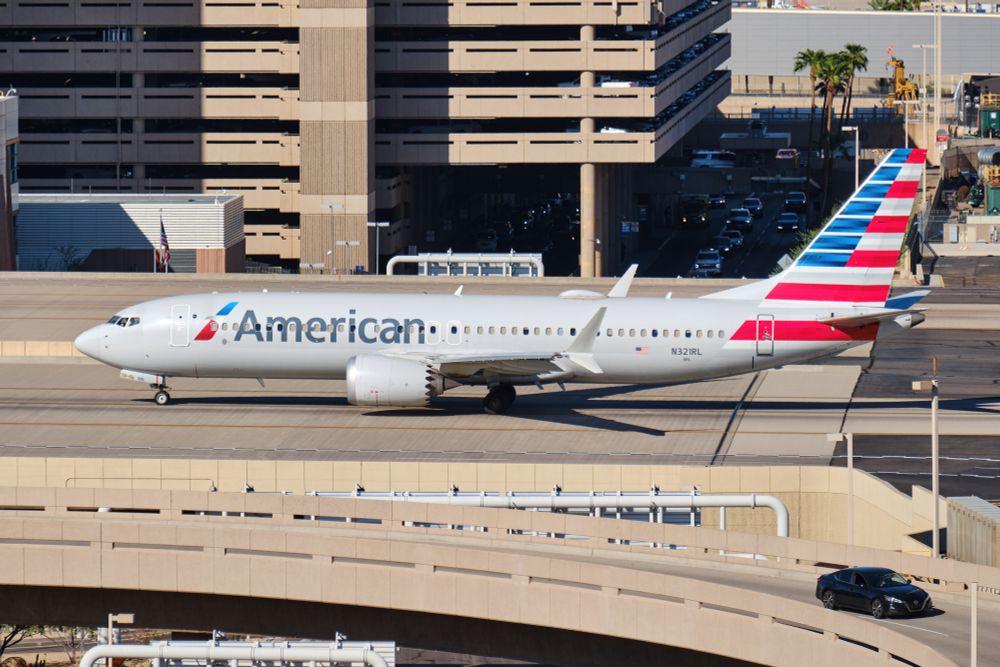 An American Airlines Boeing 737 MAX 8 (registration N321RL) taxis on an elevated section of taxiway at Phoenix Sky Harbor International Airport. The aircraft, painted in the airline’s silver livery with red, white, and blue tail stripes, passes by a tan-colored parking garage and airport infrastructure under bright morning sunlight.