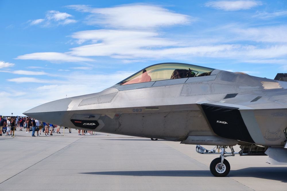 A close-up side view of the front section of a gray F-22 Raptor stealth fighter jet, parked on an asphalt tarmac under a blue sky with scattered white clouds. The cockpit canopy is visible, and the number "083" is painted on the side of the fuselage near the nose. The aircraft's front landing gear is down. In the background to the left, a crowd of people can be seen.