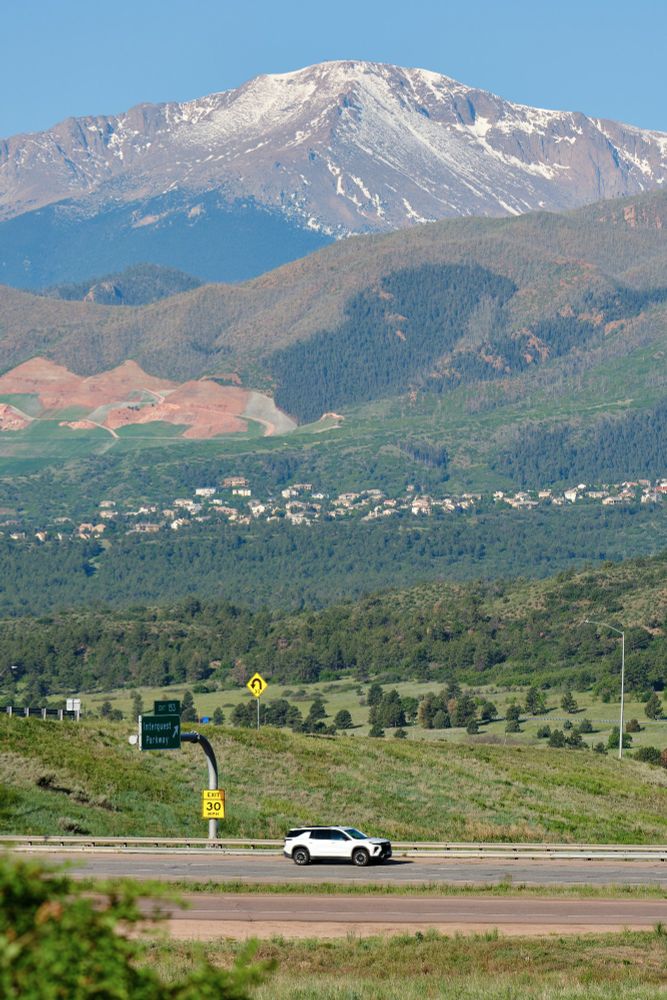     Query successful

A white SUV drives on a highway with a U-turn sign on the right and a green sign indicating "EXIT 153 Interquest Parkway" on the left. Beyond the highway, a vast expanse of green trees covers rolling hills. In the mid-ground, a residential area is visible amongst the trees, and further back, patches of reddish-brown earth are exposed on the hillsides. Towering over everything in the background is Pikes Peak, its summit still capped with snow under a clear sky.