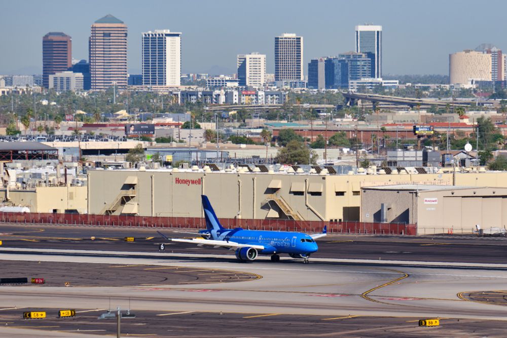 A ground-level, side-view photograph of a Breeze Airways Airbus A220-300 (N245BZ) landing at an airport. The aircraft is painted in the airline's vibrant blue livery with light blue checkmarks on the tail. In the foreground are the airport's taxiways and signage. Behind the aircraft is a large, beige Honeywell aerospace facility, and further in the distance, a portion of the dense skyline of downtown Phoenix, Arizona rises against a hazy blue sky on a sunny day.