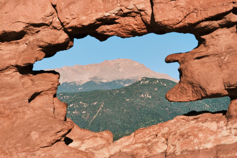 A horizontal photo of a large rock formation in the foreground with a hole that frames a view of Pikes Peak in the background. The rock formation is red and takes up most of the left and right sides of the photo, with a jagged hole in the center. Through the hole, Pikes Peak is visible with its grey, rocky summit, and below that, a large expanse of green pine trees. The sky above Pikes Peak is clear and light blue.