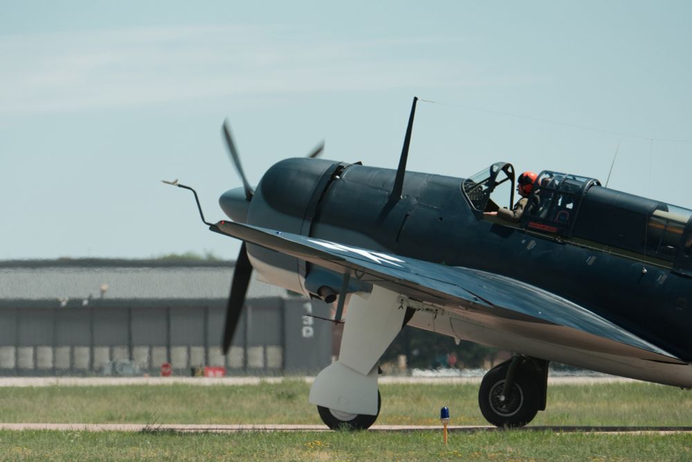 A dark blue historical military aircraft, possibly a dive bomber, with white undercarriage and a large propeller, sits on a grassy airfield. The pilot, wearing a red helmet, is visible in the open cockpit. The plane has a white star insignia on its wing. In the background, a large hangar-like building is partially obscured by the plane. The sky is light blue with some white clouds.