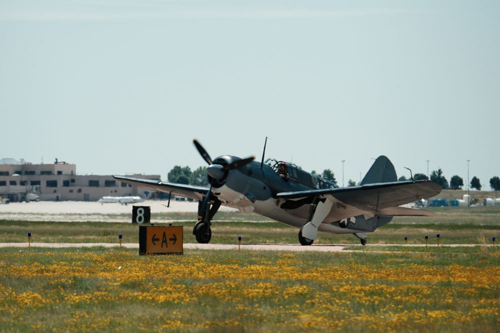 A full shot shows a SB2C Helldiver aircraft on a taxiway. The grass in the foreground is covered with yellow flowers, with buildings in the background. The aircraft is painted in a two-tone blue and white camouflage, with the white on the underside. The US white star insignia is visible on the side of the fuselage. A yellow and black sign with an arrow pointing right and the letter "A" is visible in front of the plane.