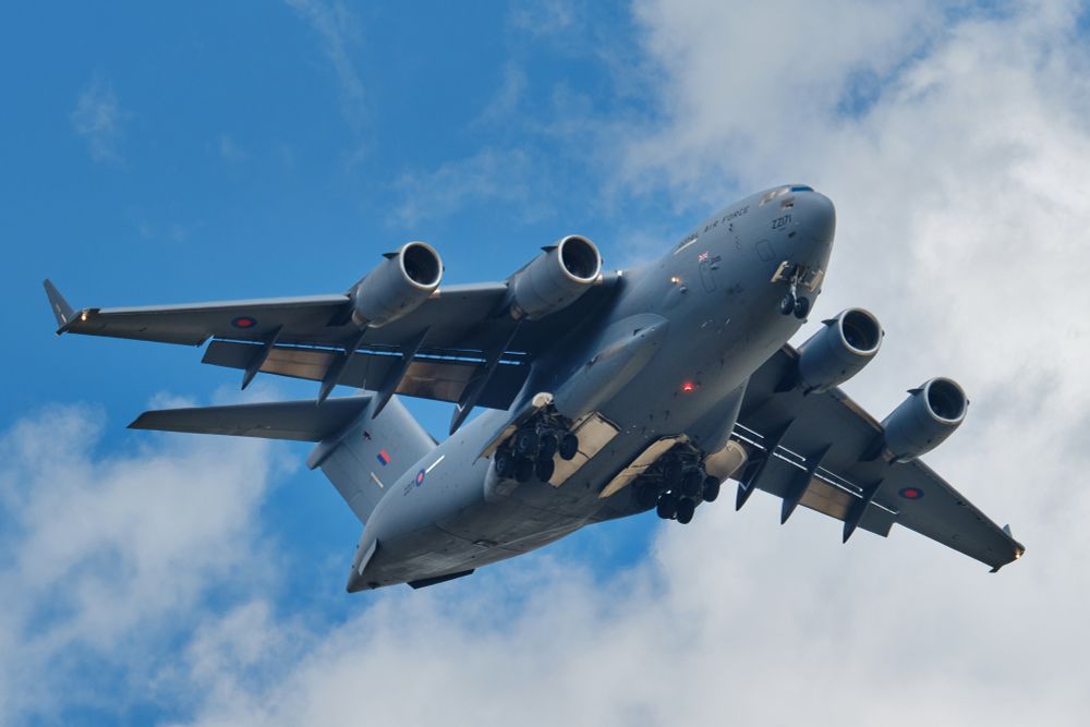 A wide, low-angle shot of a Royal Air Force C-17 Globemaster III military transport aircraft, registration ZZ171, in flight against a bright blue sky with scattered white clouds. The aircraft's landing gear is deployed, and all four engines are visible. The underside shows open gear bays and the large wings.