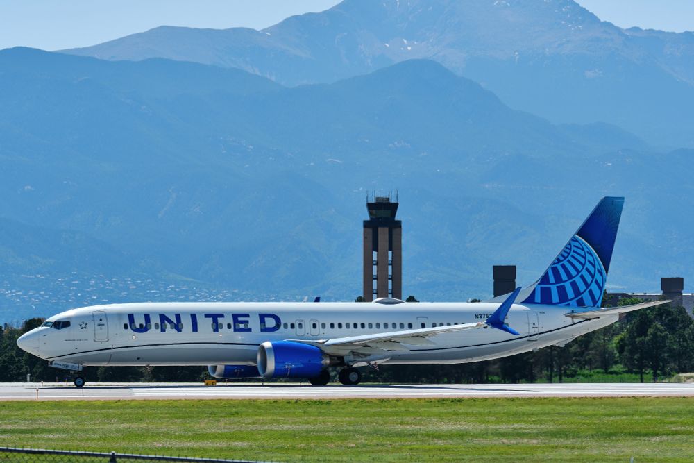 A United Airlines Boeing 737 MAX 9, registration N37536, is seen from its right side on a runway, with the large "UNITED" logo clearly visible along its fuselage. In the background, a distinctive brown air traffic control tower stands tall, and behind it large, hazy Pikes Peak rises under a smoky sky. Green grass is visible in the foreground.