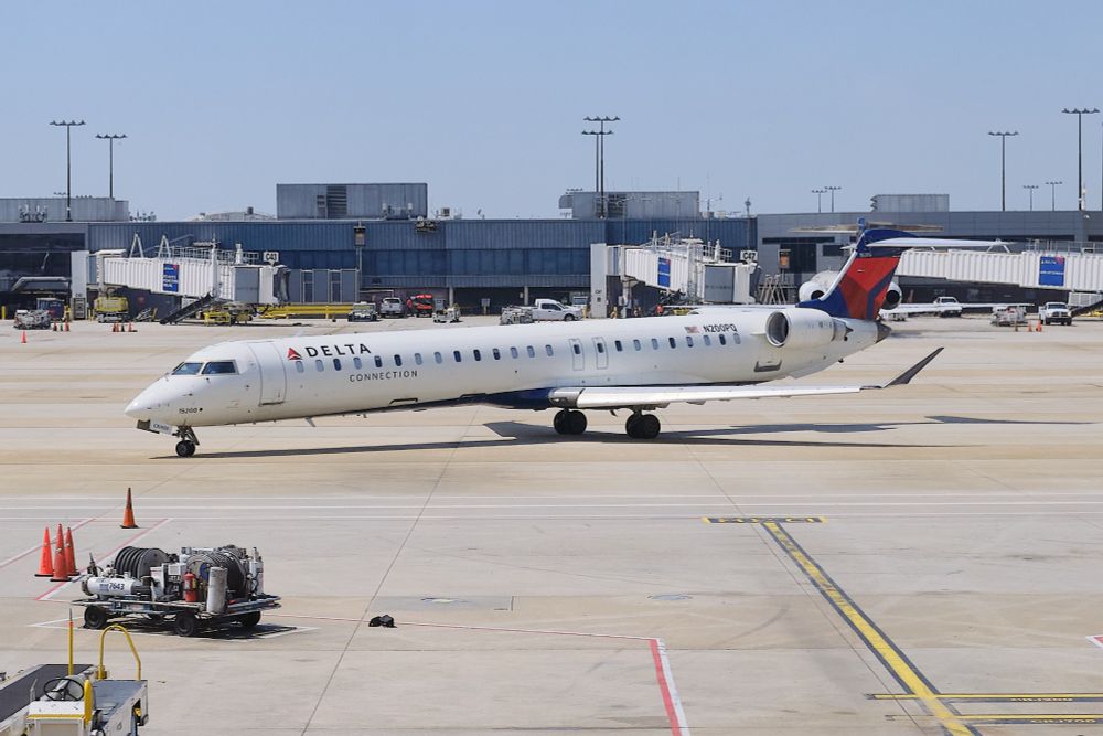 A white Delta Connection CRJ-900 regional jet, tail number N200PQ, is parked on an airport tarmac under a clear sky. The aircraft has the Delta Connection logo on its fuselage and the distinctive red and blue Delta tail livery. In the background, several jet bridges are visible connecting to airport terminals, with other ground support equipment and vehicles scattered around. The scene suggests a sunny day at a bustling airport.