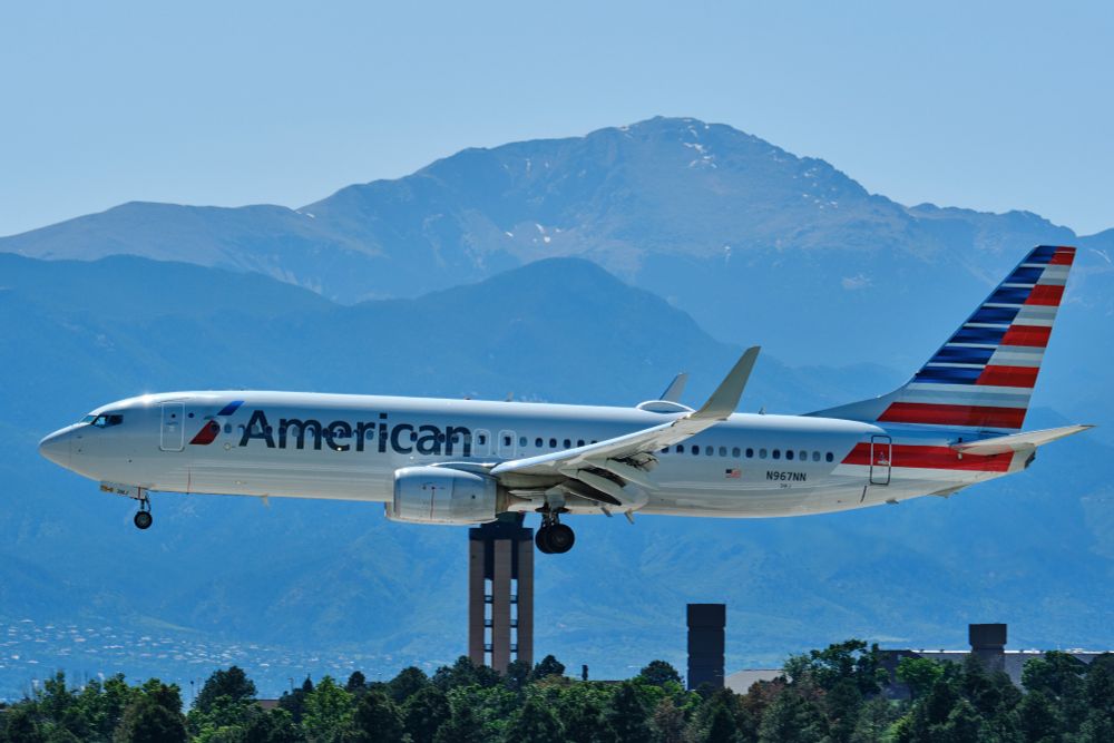 A Boeing 737 airplane, operated by American Airlines, is captured in flight against a clear blue sky. The aircraft, with registration number N967NN, displays the airline's distinctive red, white, and blue tail livery and "American" emblazoned on its fuselage. Its landing gear and flaps are deployed. Pikes Peak and the airport control tower are visible in the background.