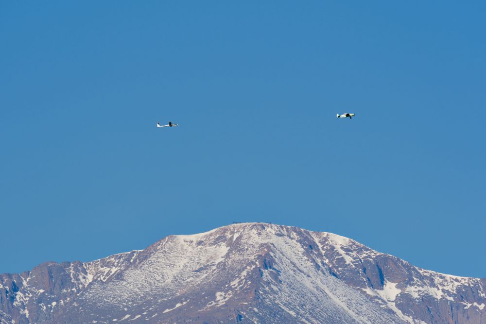 A white DG-1000S glider being towed by a tow plane to the right across a clear blue sky with the snow-capped summit of Pikes Peak below them.