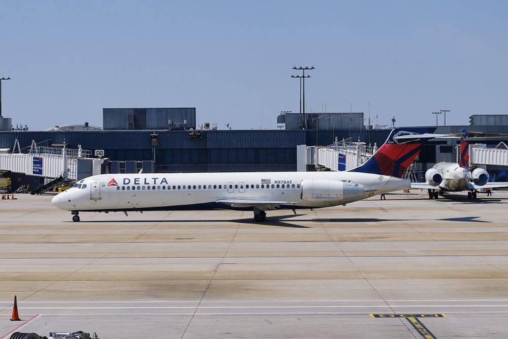 A Delta Air Lines Boeing 717 aircraft, tail number N978AT. The plane is white with the Delta logo and name prominently displayed on the fuselage. The tail features Delta's red and blue livery. To the left, a jet bridge is visible. In the background, there are other airport structures, including more jet bridges and buildings. Another aircraft, partially visible, is parked behind the featured Delta plane.