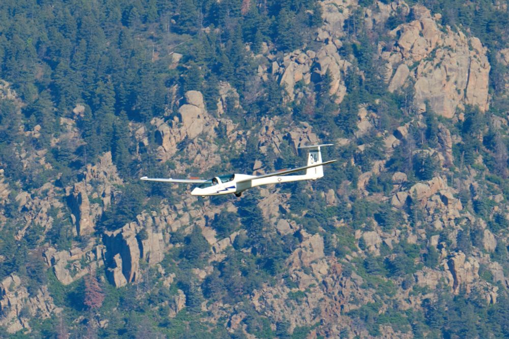 A white DG-1000S glider, seen from the side, flies in front of a densely forested, rocky mountainside.