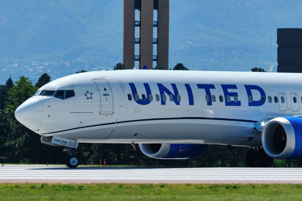 A close-up, side view of the front section of a United Airlines Boeing 737 MAX, registration N37536, turning on to a runway. The "UNITED" logo and a "Star Alliance" logo are clearly visible on the white fuselage, with blue accents on the belly and engine. In the background, part of a tall, brown control tower is visible, with hazy blue mountains and trees further in the distance under a bright sky.