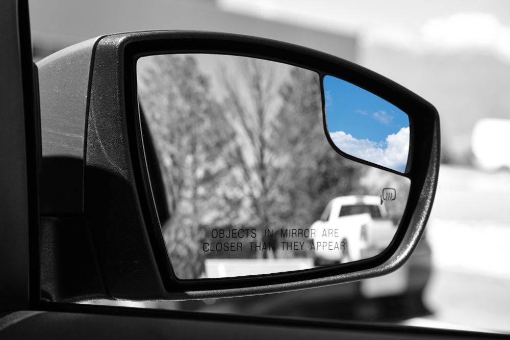 A monochrme picture of a car's side mirror, with the main mirror showing a black and white reflection of a white pickup truck, and a smaller, upper-right convex mirror showing a blue sky with white clouds in color. The main mirror has the etched text "OBJECTS IN MIRROR ARE CLOSER THAN THEY APPEAR" and a heating element symbol.