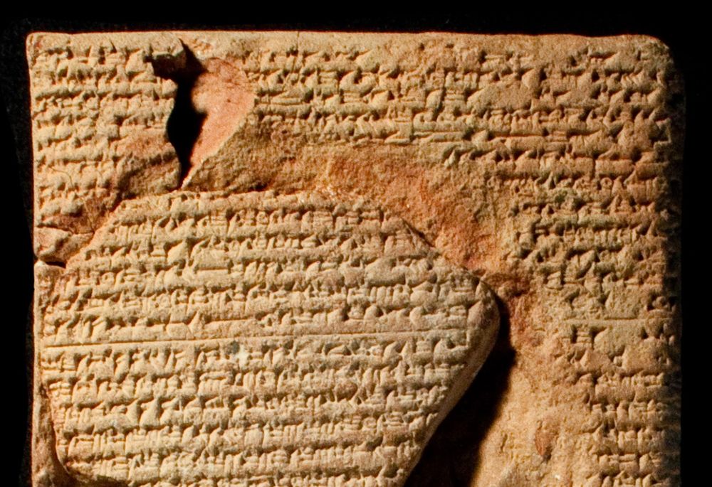 Close up of the top part of a broken, beige coloured cuneiform tablet. There are about twenty lines of text visible, and several large cracks. The background of the photo is black
