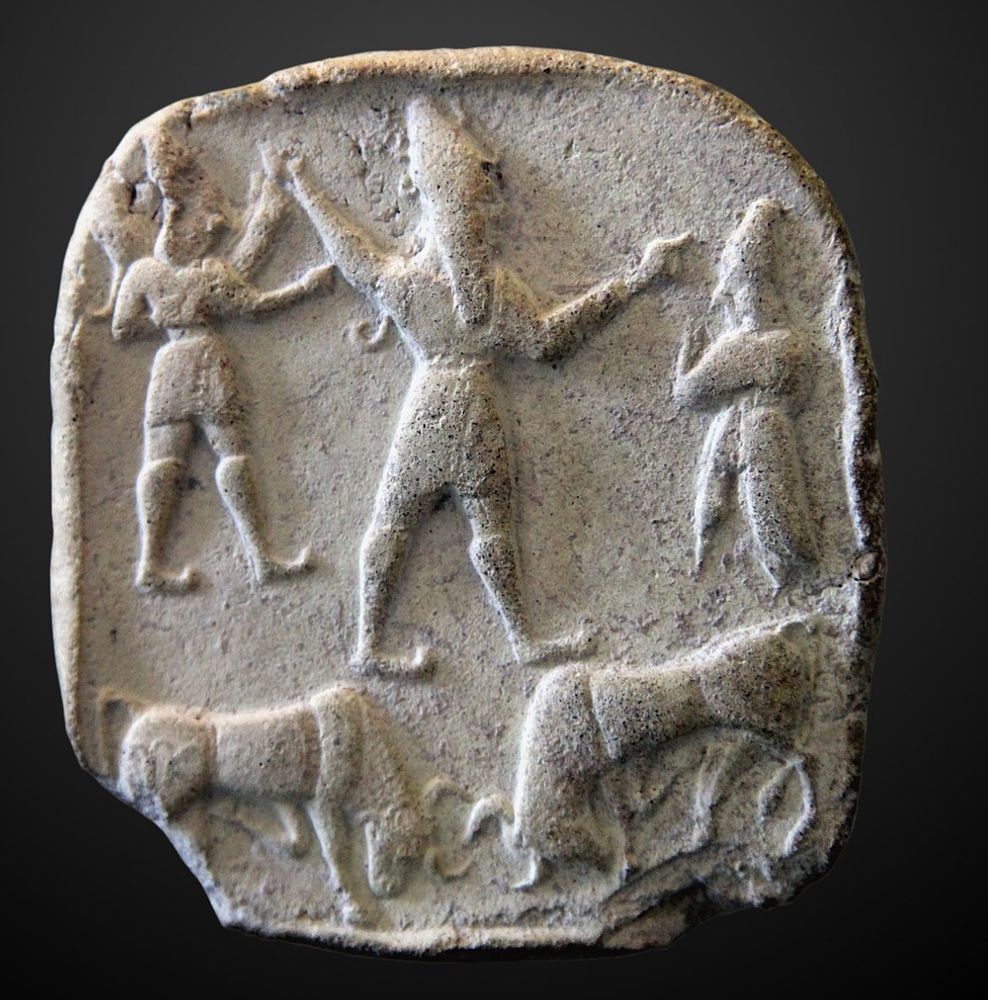 Photo of a terracotta plaque showing the Hittite storm god in the centre in profile wearing a conical hat. He is flanked by two other creatures and stands atop two bulls whose horns are locked
