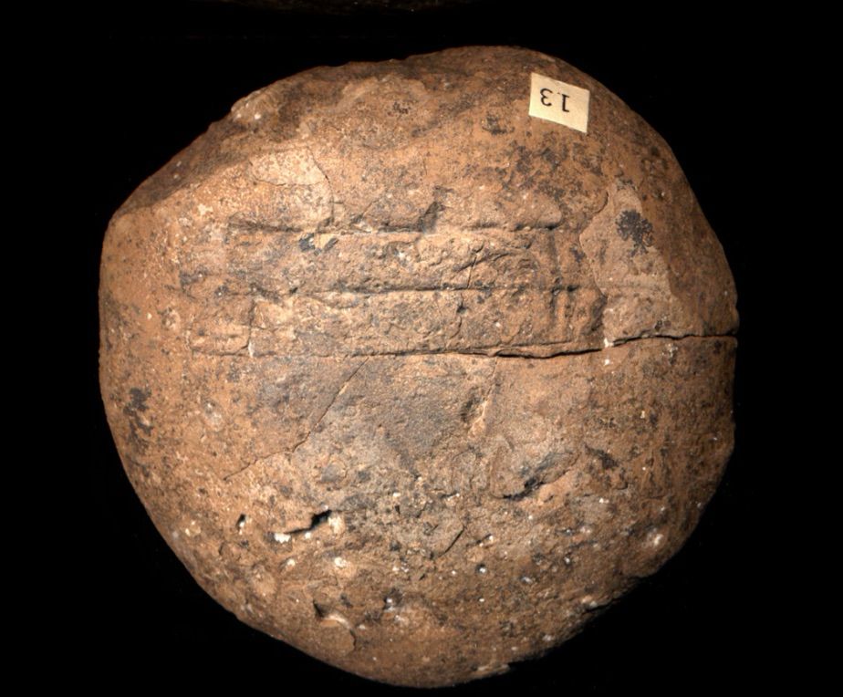 Photo of the back of a round tablet with two lines of cuneiform text and only a few signs visible. There is a sticker with the number 13 upside down at the top right of the circle. 