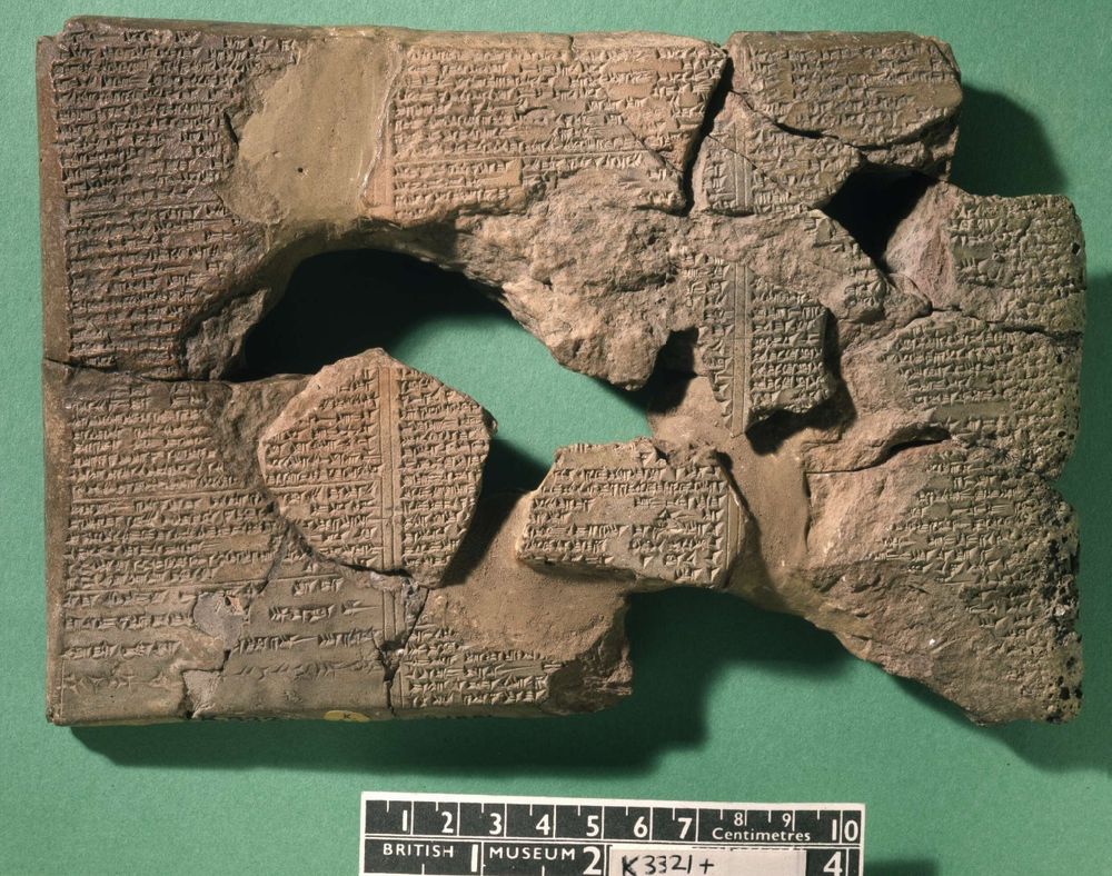 Photo of several clay tablet fragments pieced back together with several gaps throughout. The tablet is covered in cuneiform signs and is on a green table or other background. At the bottom of the image is a rule with British Museum written on it and one tablet identifier K 3321+. It shows the tablet is about 20cm wide