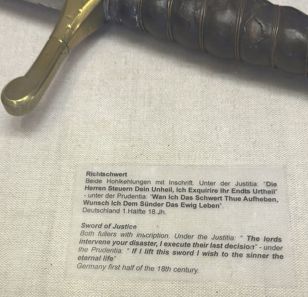Photo of a sword labeled with inscription translated from German: "The lords intervene [in] your disaster, I execute their last decision" and "If I lift this sword I wish to the sinner the eternal life".