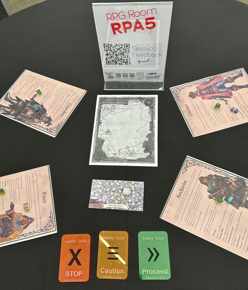 A table set up for a 4-player game of Mappa Mundi. In the centre of the table is a black & white map of the game region Barbecia. On either side are colour printouts of the player Licenses with a d4 and d6 polyhedral dice atop each one. Below the map is a card printed with a photo of seashells and the words “New Balneaire” in a banner. Below that card is a set of acrylic safety tools: red stop, orange caution, green proceed. Above the map is a sign labelling the table for the convention. 