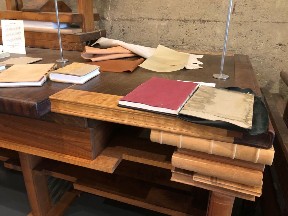 A display at the American Bookbinders Museum. Several books in various states of completion sit on a wood desk that is carved to resemble stacks of hardcover books. The book in the foreground has a red endsheet that is being prepared to glue on to the inside back cover. In the background are components for binding a book, including leather and parchment. 