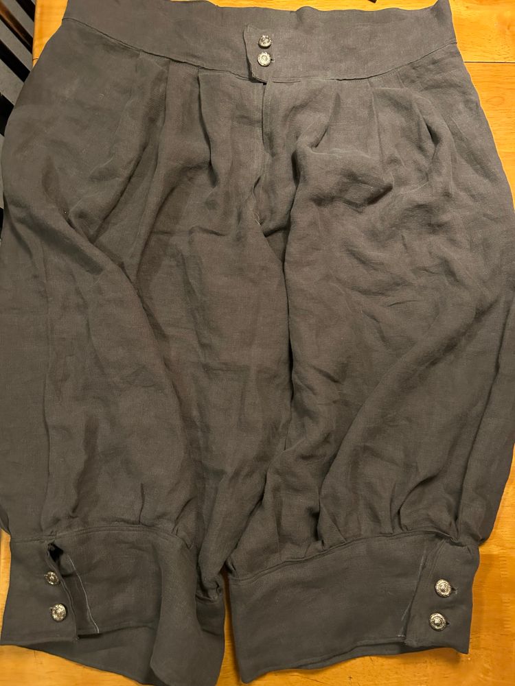 A pair of olive-green pleated, full knee-length pants in a “medieval” style with gold buttons at the waist and leg bands lying on a wood table.