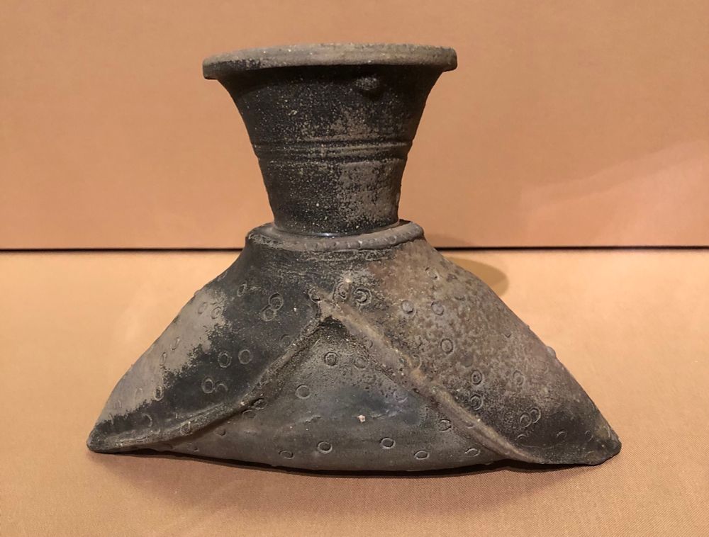 Photo of a sue ware (stoneware) piece from the Asian Art Museum in San Francisco called “Bottle in the shape of a leather bag”. The base of the piece is roughly triangular, opening to a slightly flared neck. The overall colour is a muted black, worn in places to grey or beige - especially on the sides. The clay has been imprinted to mimic the details and wear of a leather bag. 