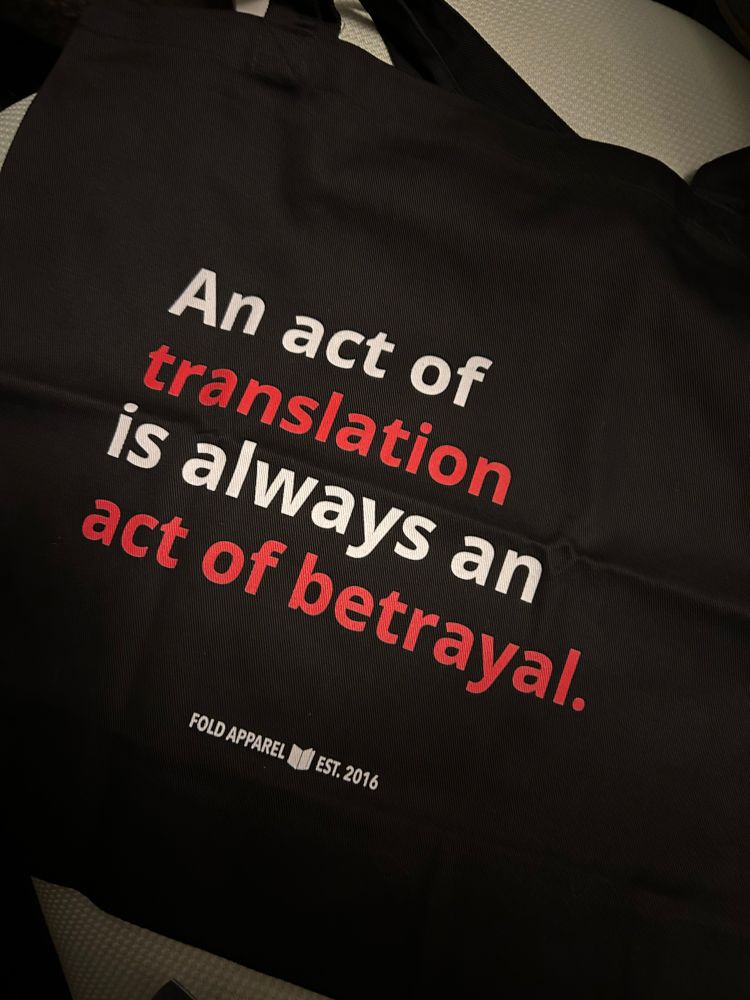 Black tote bag from Fold Apparel featuring a quote from R. F. Kuang’s novel “Babel” reading “An act of translation is always an act of betrayal.” The text is bold white with the words “translation” and “act of betrayal” coloured red.