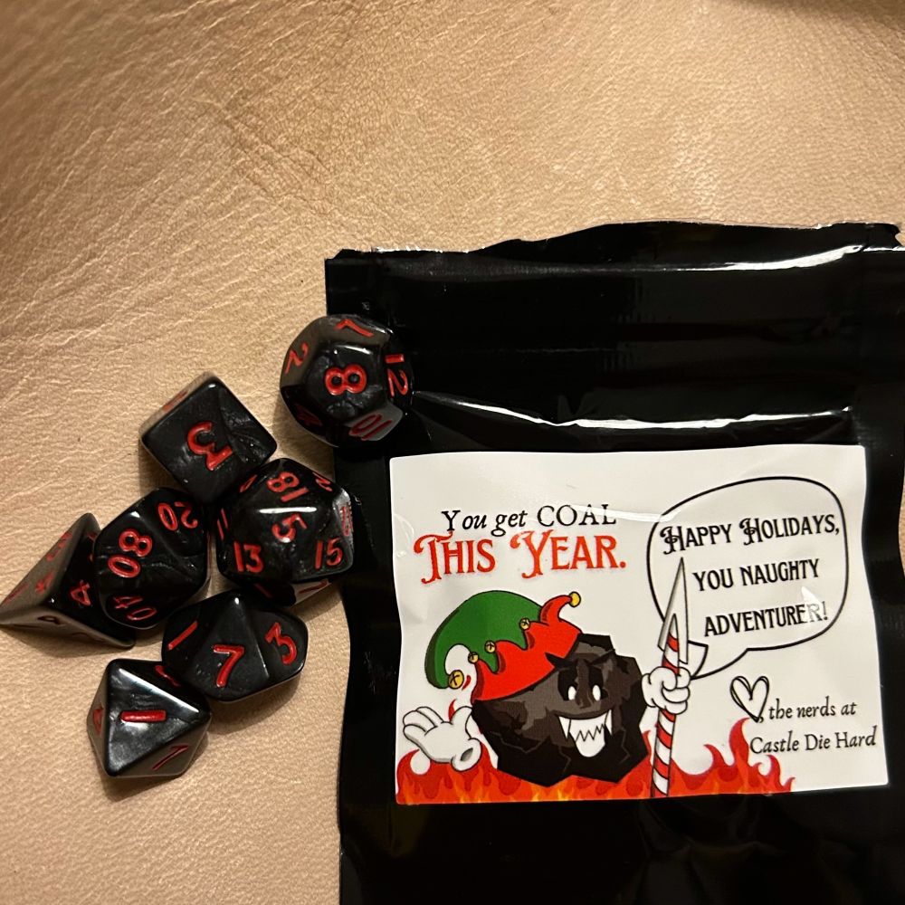 A set of 7 mini polyhedral dice in a pale leather dice tray. The dice are opaque black with shimmery silver swirls and the numbers printed in red. To the right of the dice is an opaque black bag. The bag has a white sticker on it with a drawing of an anthropomorphic lump of coal wearing a green and red holiday elf hat and white gloves, holding a candy cane striped spear. There are flames along the bottom of the drawing. There is a speech bubble for the coal saying “Happy holidays, you naughty adventurer!”. The text on the sticker reads “You get coal this year. ❤️the nerds at Castle Die Hard”
